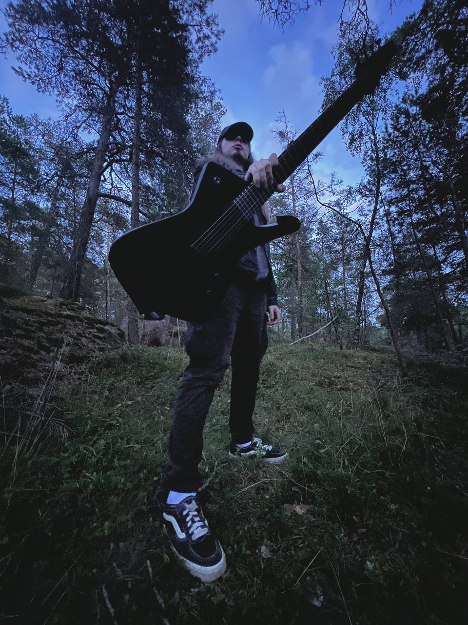 Standing in a pine forest at dusk holding a seven-string electric guitar